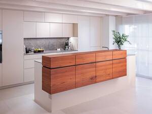 Die Küche «Nussbaum Theke» ist wie alle Projekte von Schneebeli individuell und massgefertigt auf die Wünsche der Kundschaft. Das Nussbaumholz-Element fungiert hier nicht nur als Treffpunkt für Alltag und Genuss und als eleganter Blickfang, sondern es reduziert auch das Sichtfeld auf den Koch- und Spülbereich. Schneebeli AG.