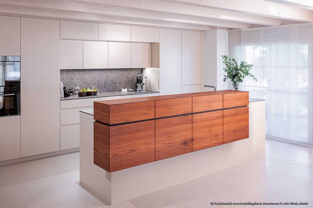 Die Küche «Nussbaum Theke» ist wie alle Projekte von Schneebeli individuell und massgefertigt auf die Wünsche der Kundschaft. Das Nussbaumholz-Element fungiert hier nicht nur als Treffpunkt für Alltag und Genuss und als eleganter Blickfang, sondern es reduziert auch das Sichtfeld auf den Koch- und Spülbereich. Schneebeli AG.